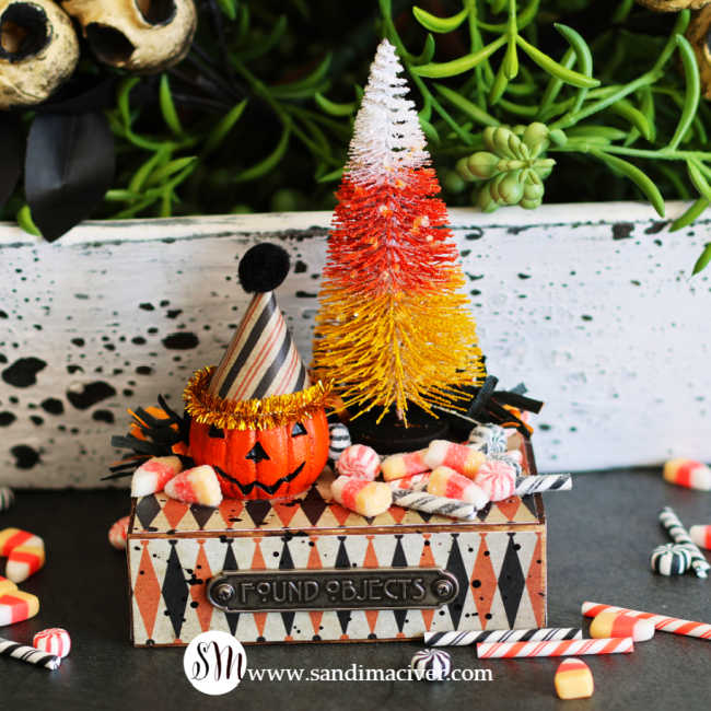 Tim Holtz Halloween vignette featuring a jack-o’-lantern in a striped party hat, a candy corn-colored bottle brush tree, and miniature candies on a patterned pedestal with a Found Objects label.