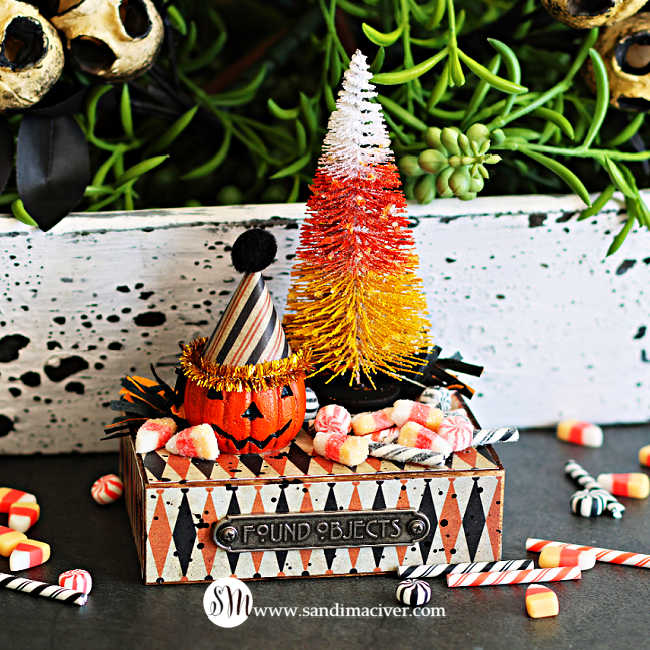 Tim Holtz Halloween vignette featuring a candy corn-colored bottle brush tree, a jack-o’-lantern wearing a striped party hat, and glittery miniature candies arranged on a vintage-style pedestal with a Found Objects plaque.