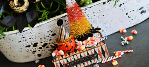 Tim Holtz Halloween vignette featuring a jack-o’-lantern in a party hat, a colorful candy corn bottle brush tree, and miniature candies displayed on a decorated pedestal.