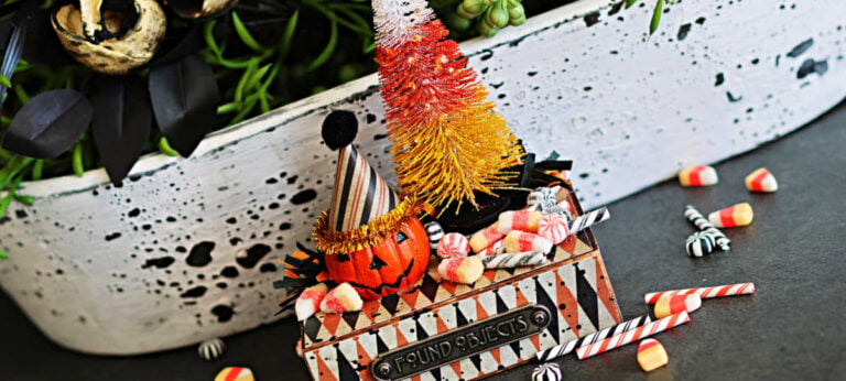 Tim Holtz Halloween vignette featuring a jack-o’-lantern in a party hat, a colorful candy corn bottle brush tree, and miniature candies displayed on a decorated pedestal.