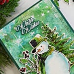 Messy watercolor Christmas card featuring a snowman and evergreen trees on a green and aqua background with white snowflakes and festive greenery.