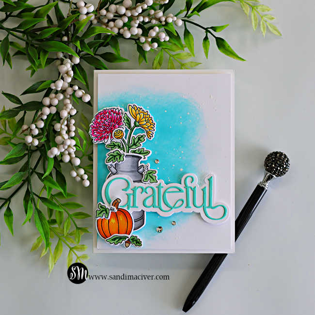 Handmade fall card featuring colorful mums and a pumpkin arranged around a watering can, with a large Grateful sentiment over a turquoise ink-blended background.