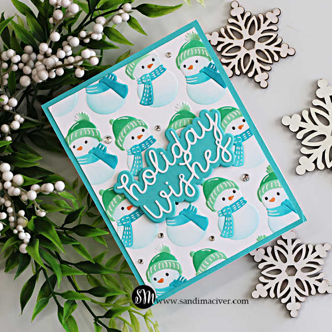 Cozy Snowman embossed and stenciled Christmas card with teal and green snowmen, a dimensional Holiday Wishes sentiment, sparkling gem accents, greenery, and wooden snowflakes.
