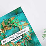 Handmade Christmas card featuring a gold Leafy Wreath over a vibrant teal and green background created with Lindy’s Magicals, accented with gold flowers, gems, and a “May Your Christmas Be Merry” sentiment.