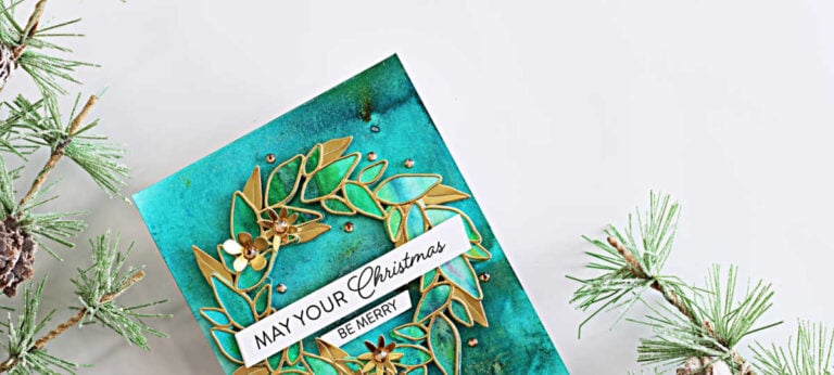 Handmade Christmas card featuring a gold Leafy Wreath over a vibrant teal and green background created with Lindy’s Magicals, accented with gold flowers, gems, and a “May Your Christmas Be Merry” sentiment.