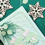 Mint Stitched Mittens Christmas card with white cuffs, layered die-cut greenery, a Seafoam embossed background and frame, and a “Be Merry and Bright” sentiment.