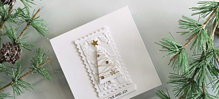 White-on-white Christmas card featuring a Simple Trees die-cut wrapped in gold thread, gold gems and stars, an embossed botanical panel, a postage-edge frame, and a “Love and Joy” sentiment.