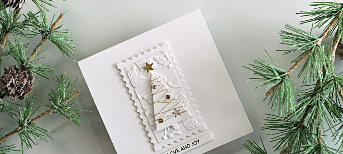 White-on-white Christmas card featuring a Simple Trees die-cut wrapped in gold thread, gold gems and stars, an embossed botanical panel, a postage-edge frame, and a “Love and Joy” sentiment.