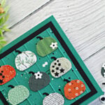 Handmade Thanksgiving card featuring nine patterned die-cut pumpkins on a teal embossed wooden plank background with black matting, including a pumpkin with repeating “thankful for you” text, photographed beside greenery and extra pumpkin die cuts.