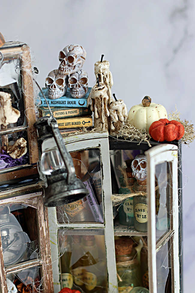 Close-up of the right side of Tim Holtz Collector’s Laboratory, featuring miniature books topped with skulls, dripping candles, pumpkins, and distressed glass-front cabinets filled with potion bottles.