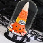Tim Holtz Trick or Tree Halloween display featuring a candy corn-colored bottlebrush tree atop a jack-o’-lantern, miniature candies, and a numbered plaque under a glass dome tied with a black-and-white bow.