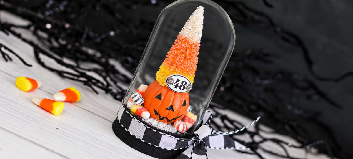 Tim Holtz Trick or Tree Halloween display featuring a candy corn-colored bottlebrush tree atop a jack-o’-lantern, miniature candies, and a numbered plaque under a glass dome tied with a black-and-white bow.