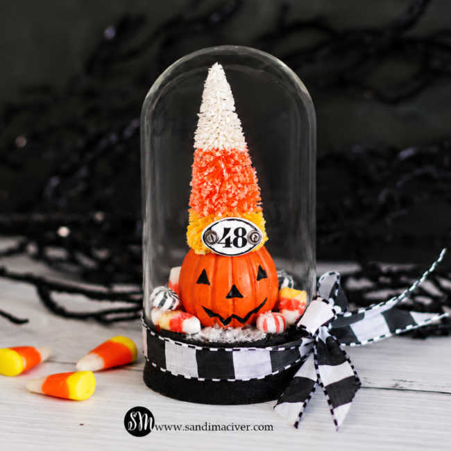 Front view of Tim Holtz Trick or Tree Halloween decor featuring a candy corn-colored bottlebrush tree, jack-o’-lantern, “48” plaque, and miniature candies inside a glass dome with a black-and-white ribbon bow.