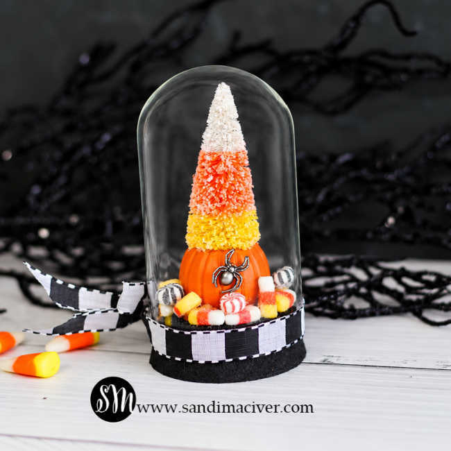 Back view of Tim Holtz Trick or Tree Halloween decor featuring a candy corn-colored bottlebrush tree atop an orange pumpkin with a silver spider, surrounded by miniature candies under a glass dome trimmed with black-and-white ribbon.